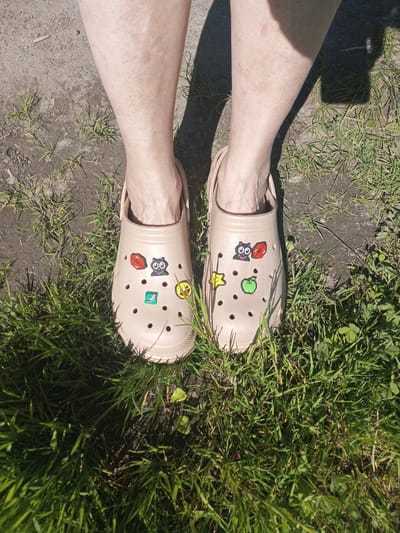 The image provides a top-down view of a person's lower legs and feet, clad in light beige Crocs clogs, standing on a patch of ground. The setting is outdoors, characterized by sparse green grass interspersed with patches of dry dirt or concrete.
Bright, direct sunlight is evident from the distinct, dark shadows cast by the legs and footwear onto the ground, indicating a clear and sunny day, likely during midday or early afternoon. The person's bare legs, visible from mid-calf down to the ankles, display fair skin with some visible freckles or minor blemishes.
The beige Crocs are customized with several colorful, pixelated Jibbitz charms. The left clog features a red pixelated mouth, a black pixelated cat with green eyes and a pink nose, a green pixelated square icon (resembling a message or paper symbol), and a yellow pixelated smiley face with a sticking-out tongue. The right clog is adorned with a black pixelated cat with green eyes, a red pixelated mouth, a yellow pixelated star, and a green pixelated apple.
The scene captures a static moment, showcasing the footwear and the immediate outdoor environment in Kommunar, Russia. No specific activity or emotion is discernible, and there is no visible text within the image.