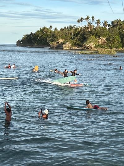 The image depicts a coastal scene in General Luna, Philippines. A group of people are surfing in the water. There are surfboards of different sizes and colors visible. One person appears to be falling off a surfboard, while others are attempting to balance or stand. In the background, a green landmass with trees, including palm trees, is visible. The water is blue and choppy. Some individuals are swimming. The sky is partly cloudy.