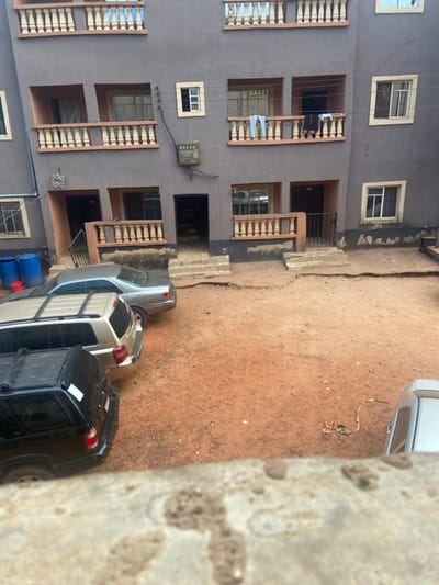 This image depicts a residential compound in Nsukka, Nigeria, viewed from an elevated perspective. The central subject is a three-story building with a gray plastered facade.
The building features multiple balconies on its first and second floors, each equipped with ornate, light-colored concrete balustrades. Rectangular windows with white frames are visible across all floors. Ground-level entrances, some fortified with black metal security gates, lead into the building. A utility meter box is mounted on the first-floor wall, positioned between two balconies. Light-colored trousers and dark garments are observed hanging to dry over the balustrades of two first-floor balconies.
The foreground and courtyard consist of unpaved, reddish-orange laterite soil, characteristic of the region, which appears uneven and somewhat barren. Three vehicles are parked consecutively on the left side of the unpaved area: a silver sedan, followed by a beige SUV, and a black SUV, all facing left. The rear section of a fourth silver vehicle is partially visible in the lower right corner of the frame. Near the left side of the building, two blue plastic drums and one red bucket are present on the ground. No individuals are visible within the scene. The image is taken during daylight hours.