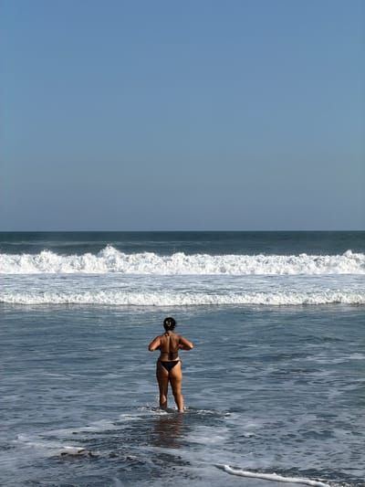 The image captures a serene ocean view in Selemadeg, Indonesia. An adult female, viewed from behind, stands in the shallow water with foam and ripples around her lower legs. She wears a black two-piece swimsuit. Her dark hair is pulled up into a bun at the back of her head. Her arms are bent at the elbows, with her hands positioned in front of her body as she looks out towards the sea. The sky above is a clear, uniform light blue, meeting a distinct horizon line that separates it from the ocean. In the mid-ground, multiple lines of white-capped waves break, with a prominent wave cresting across the scene. The water further out is a darker blue-grey and appears calmer.