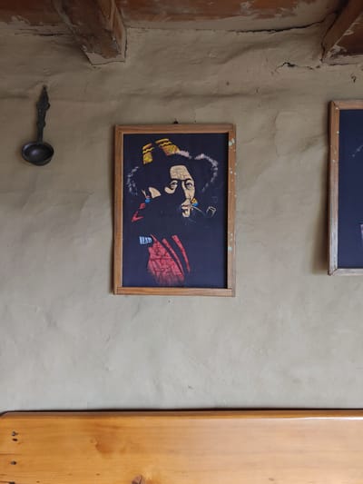 The image depicts an interior scene with a light-colored, textured wall that shows signs of cracking and wear. A wooden beam is visible near the ceiling, contributing to a rustic aesthetic. To the left, a dark metal ladle with an ornate handle hangs on the wall. In the center of the frame, a framed artwork hangs. The artwork is a colorful depiction of a person, possibly Bob Marley, with a pipe in their mouth and wearing a fur-lined hat and a red garment. To the right, another framed object, appearing to be a dark blackboard or framed picture, is partially visible. Below the wall and artwork, a smooth, light-colored wooden bench or ledge spans the width of the frame, showing visible nails and wood grain. The scene appears to be in a building with a natural, unfinished material for the walls and ceiling, suggesting a location like Prini, India, where such architectural styles are common in guesthouses or traditional homes.