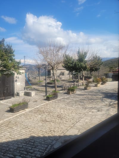 The scene depicts a sunny outdoor area in Κωστήτσι, Greece. The foreground features light-colored stone paving, a combination of flagstones and cobblestones, with a prominent shadow cast from the lower right. Several dark wooden rectangular planters, each containing green foliage, are placed on this paving.
The mid-ground centers on a village square. To the left, a traditional stone building with a large dark wooden door stands next to a stone utility structure displaying a partially legible sign and a small bell. A blue conduit or hose runs down its wall. The square is furnished with multiple dark wooden benches and black, multi-lantern streetlights. Several trees are present; some are deciduous and bare, while others are evergreen. A metal railing borders the far side of the square, offering a view towards distant hills or valleys.
The background consists of green, tree-covered mountains beneath a bright blue sky dotted with scattered white cumulus clouds. No individuals are visible within the depicted environment.