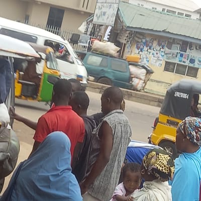In Dadi Riba, Nigeria, the image shows individuals gathered near vehicles on a street. People are loading or unloading items. A yellow tricycle is visible. Buildings are present along the street. Luggage is on the ground.