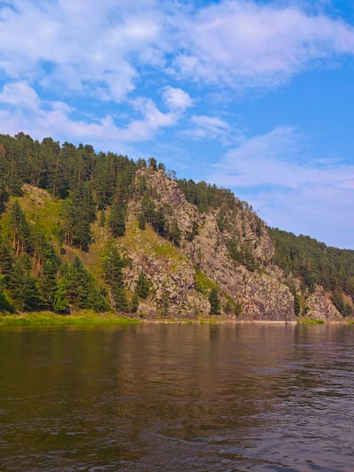 The image depicts a wide, tranquil river flowing in front of a steep, wooded mountainside. The mountain is covered with a dense forest of evergreen trees, primarily pines, with patches of rocky outcrops and green vegetation interspersed throughout. The river's surface reflects the colors of the sky and the surrounding landscape, creating a shimmering, slightly distorted view of the mountain and trees. The sky above is a clear blue with scattered white clouds, suggesting fair weather. The scene conveys a sense of natural beauty and serenity, characteristic of a remote, unspoiled natural environment. This scene is likely located in the natural landscapes surrounding Beregovaya-Podemnaya, Russia, a region known for its picturesque river valleys and forested mountains.