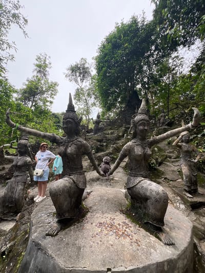 In the heart of Ko Samui, Thailand, a surreal landscape unfolds at the Secret Buddha Garden, also known as the Tarnim Magic Garden. The scene is dominated by weathered, ancient-looking statues depicting mythical beings and figures from local folklore. Two prominent statues, gracefully posed with outstretched arms, anchor the foreground. Their intricate details, though softened by time and the elements, suggest a rich cultural heritage. Between them, a small, carved creature, possibly a monkey, sits, adding a touch of whimsy to the solemn tableau. Scattered around the base of the statues are numerous coins, likely offerings from visitors seeking blessings or paying respects.

The figures are set amidst a lush, verdant jungle, with trees and foliage creating a natural canopy overhead. The ground is a rocky, uneven terrain, characteristic of the island's natural topography. In the background, more statues are visible, suggesting a sprawling garden filled with these unique sculptures.

Three individuals are present in the image. A woman, wearing a straw hat, sunglasses, a white t-shirt with a butterfly motif, and light blue shorts, stands to the left of the main statues, her hands on her hips, observing the scene. Beside her, a child in a light blue hoodie and shorts is also present. Another person, partially obscured, can be seen further back in the garden, adding to the sense of exploration and discovery within this unusual place. The overall atmosphere is one of quiet contemplation, where art, nature, and spirituality converge.