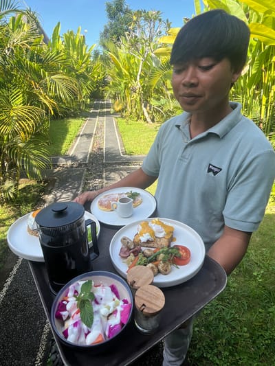 In Tegallalang, Indonesia, a man in a light blue polo shirt with a "Wrangler" logo on the left chest is holding a dark gray tray with several plates of food. In the foreground, there is a bowl of fruit salad topped with mint. To its right are two small wooden-lidded jars. Further back on the tray, a black French press sits next to a plate with a fried egg and some salad. Another plate has two pancakes with syrup and a small pitcher of syrup. A third plate features a meal with eggs benedict, vegetables, and a sliced tomato. The man is standing on a path lined with dark gray stones and decorative white pebbles, surrounded by lush green foliage and palm trees. The sky above is clear and blue.