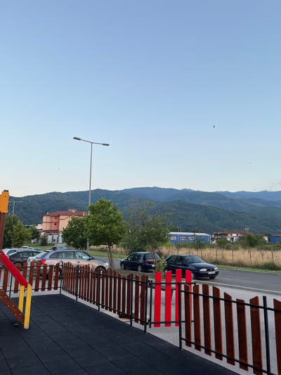 The image depicts a view of a street and distant mountains, likely in or near Petrich, Bulgaria. In the foreground, a playground area with black rubber matting and a wooden fence with red accents is visible on the left. Beyond the fence, several cars are parked along the side of a road, including a silver station wagon, a dark blue hatchback, and a dark blue sedan. A tall street lamp stands on the right side of the road. In the middle ground, buildings and trees are present, including a multi-story building with an orange facade. The background is dominated by rolling, tree-covered mountains under a clear blue sky. A bird is seen flying in the sky. The scene appears to be set in the late afternoon or early evening, with soft lighting.