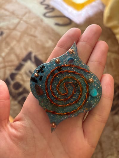 A person's hand holds a blue, glittery resin pendant shaped like an elongated hexagon with two points extending from one side. The pendant is embedded with various objects, including small dark stones, copper-colored beads, a spiral of copper wire, and a small turquoise stone. The background is a blurred, patterned fabric in shades of brown and yellow.