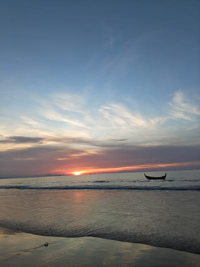 This is a vertical, eye-level, medium shot of a sunset over the ocean in Curehtunong, Indonesia. The sky is a gradient of blue and orange, with wispy clouds scattered across it. The sun is a bright orange disc, low on the horizon, casting a golden glow on the water. A single, dark boat with a curved hull floats on the calm sea. Gentle waves lap at the sandy shore in the foreground, reflecting the colors of the sunset. The wet sand also shows reflections and a small, translucent object, possibly a shell or piece of plastic, sits near the water's edge. In the distance, the faint silhouette of landmass is visible. There are no people visible in the scene.