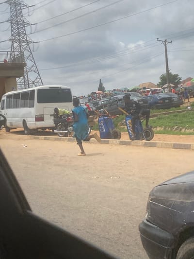 This media file captures a bustling street scene in Kuchingoro, Nigeria, under an overcast daytime sky. The image shows a roadside with a mix of vehicles, pedestrians, and commercial activity.
In the foreground and mid-ground, the scene is dominated by a concrete or asphalt road. On the left side of the road, several cars are parked bumper-to-bumper along a grassy shoulder, behind which numerous people are gathered around what appear to be market stalls or makeshift vendor setups, many featuring colorful umbrellas.
Several individuals are visible on the road. Most prominently, two people are pushing blue, two-wheeled carts. The cart closer to the viewer clearly displays the word "KINGSUN" in white lettering on its side. Another person, wearing a light blue garment, walks near a white mini-bus, which is either parked or moving slowly on the right side of the road. A motorcycle is also visible near the mini-bus, with a person sitting on it.
Overhead, a network of utility poles and power lines crisscrosses the sky, indicative of urban infrastructure. In the far background on the left, large metallic high-tension power line towers are visible against the cloudy sky. The overall impression is one of everyday street commerce and movement in a lively, populated area.