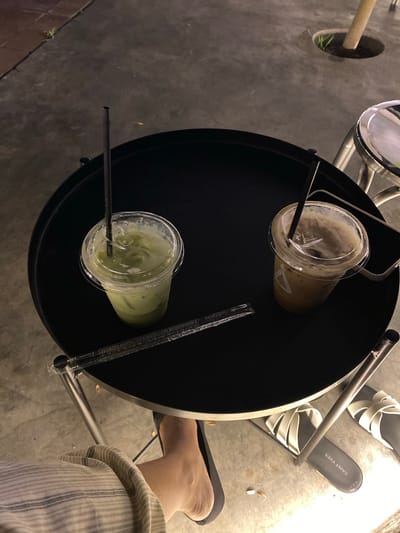 Two iced drinks, one green and one brown, sit on a black circular table. Each drink is in a clear plastic cup with a clear plastic lid and a black straw. A single black straw lies on the table next to the drinks, and a wrapper for straws is also visible. The table has a metal base and is outdoors, with a concrete ground. A person's foot, wearing a sandal, is visible at the bottom of the image. The overall lighting suggests it is nighttime or dusk. The image does not contain enough information to determine the specific location in Madiun, Indonesia.
