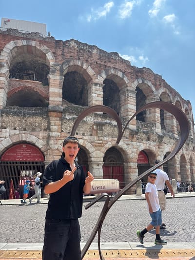 The image depicts a man standing in front of the Arena di Verona in Verona, Italy. The Arena is a large, ancient Roman amphitheater with a facade of arched openings. A modern art installation made of metal loops stands in front of the Arena. The man is wearing a black shirt and pants, a baseball cap, and pointing to the sky with both hands. Behind him, there are people walking on the sidewalk. Signs are visible, including one indicating entrance gates to the Arena and another with the word "Arena" on it. The sky is blue with some clouds.