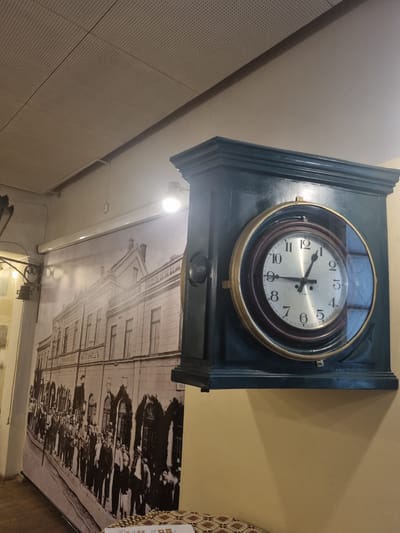 The image shows an interior setting featuring an antique clock mounted on a wall. The clock has a circular face with numbers and hands indicating a time between 9:20 and 9:30. The clock is encased in a square, dark-colored frame with decorative molding. Below the clock, a black and white image is visible. The image depicts an old building with a crowd of people in front of it. A sign on the building reads "ХАТИПЕШЪ" in Cyrillic letters. The image suggests a historical scene, likely a public gathering or event. The surroundings include parts of walls and ceilings with different textures and colors, indicating a room within a building. Given the Cyrillic inscription, the location is likely in a country that uses the Cyrillic alphabet. Based on the inscription on the wall photograph, which mentions "ГОРНА ОРЯХОВИЦА", the location is Gorna Oryahovitsa, Bulgaria.