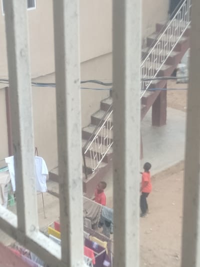 The image shows an outdoor scene behind bars. Two young boys in red shirts are visible, one standing near a staircase and the other further away. Laundry is hung on clotheslines in the foreground, with various colored clothes. A concrete staircase with a white railing leads up to a building. The ground is dirt. The setting appears to be a residential area, potentially within the city of Shangisha, Nigeria, characterized by simple architecture and a focus on outdoor living spaces. The overall impression is one of everyday life in a community.