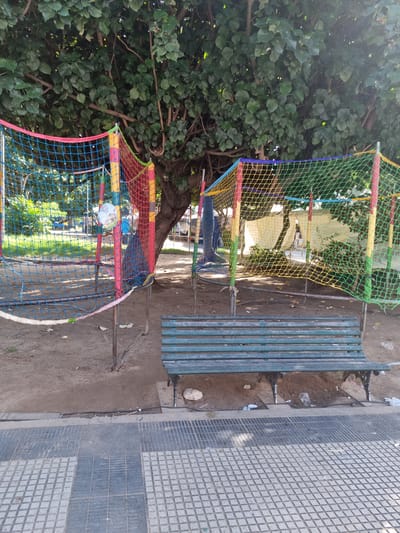 This image captures an outdoor scene, likely a public park or recreational area in Santa Cruz de la Sierra, Bolivia, observed from an elevated or tilted perspective. The setting features a large tree with dense green foliage occupying the left side of the frame, providing shade.

In the foreground and midground, a vibrant, multi-colored net structure, possibly part of a play area or a protective barrier, dominates the view. The net is composed of red, yellow, green, and blue sections, supported by various metal poles. Entangled within the net on the lower left, a partially deflated white ball or balloon, possibly depicting a cartoon character or a globe, is visible.

To the right of the net structure, a weathered park bench, painted in a faded green or blue, is positioned on a dirt ground. The ground beneath and around the bench is primarily barren earth, with some sparse dry vegetation and noticeable litter, including plastic bottles and wrappers, indicating some neglect. Adjacent to the bench and extending towards the right edge of the frame is a paved area, resembling a sidewalk, constructed with distinctive square gray tiles, some lighter and some darker, arranged in a grid pattern.

The scene appears to be devoid of people, suggesting a quiet moment. The bright, even lighting indicates it is daytime, likely a clear and sunny day, casting some soft shadows but no harsh midday glare. No specific activity or event is actively taking place. There is no visible text in the image.