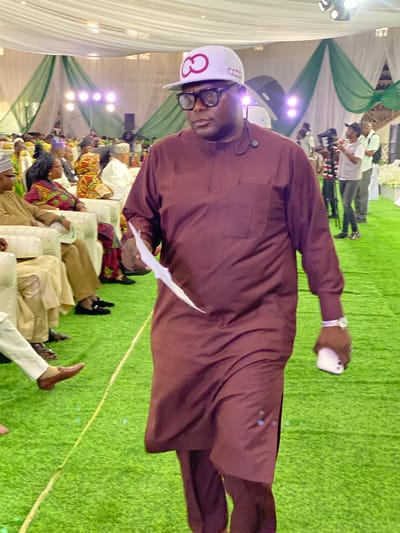 A man in a maroon tunic and pants, wearing a white baseball cap and glasses, walks across a green carpeted area. He holds a white piece of paper in his left hand and a smartphone in his right. He appears to be attending an indoor event with an audience seated on white chairs in the background. The venue is decorated with green and white draped fabric and illuminated by spotlights. The man is the primary subject, facing forward with a neutral expression. The environment suggests a formal gathering or conference.