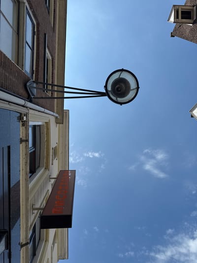 The image is an upward-facing shot of buildings and the sky, taken on a sunny day. The scene is oriented vertically. On the left, a brick building with windows is visible. Below it, a lighter colored building has a red sign with orange text that reads "TINKER.NET". A street lamp with a glass enclosure is prominently featured in the center, extending out from the brick building. The sky is blue with scattered white clouds. In the upper right corner, a portion of another building with a dormer window is visible. The image appears to be taken in the city of Deventer, Netherlands, likely in a commercial or residential area with shops on the ground floor.
