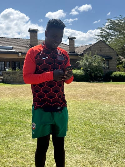 An adult male stands outdoors on a green lawn in the Olkaria ward, Kenya, under a blue sky with white clouds. The man has a dark complexion, short dark hair, and is smiling while looking down at and interacting with a dark-colored smartphone held in both hands. He wears a red and dark-patterned long-sleeved athletic top with a white Nike logo and green athletic shorts with a circular emblem on the left leg. A black waistband or small pack is visible at his waist. In the background, a two-story stone house with a reddish-brown tiled roof and multiple chimneys is visible. Two solar water heaters are on the left side of the house roof. Green shrubs and trees surround the house.
