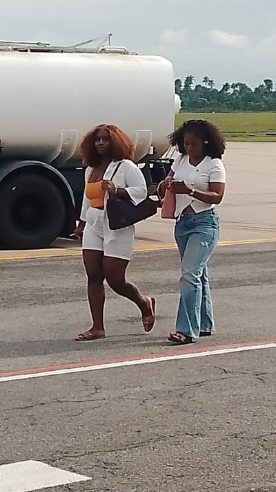 Two women are walking side-by-side on an asphalt surface, likely an airport tarmac, in Orerokpe, Nigeria. The woman on the left, with auburn hair, wears an orange crop top and white shorts with a matching white open-front shirt. She carries a dark brown handbag over her shoulder and is wearing flat, open-toed sandals. The woman on the right, with dark curly hair, is dressed in a white short-sleeved top and blue flared jeans. She is holding a pink handbag and also wears open-toed sandals. Behind the women, a large, white cylindrical vehicle, possibly a fuel tanker or a water truck, is partially visible, with its black tire prominent. The background suggests an outdoor setting with a cloudy sky, green vegetation, and palm trees. The ground is marked with yellow and red lines, indicating a designated pathway or runway.