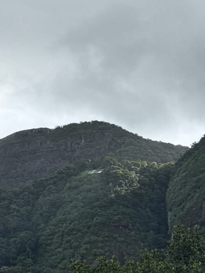 The image displays a dramatic and steep mountainous landscape under an overcast sky. The upper left portion of the frame is dominated by a light grey, cloudy sky, suggesting either an overcast day or foggy conditions.
The bulk of the image on the right is occupied by a towering mountain or cliff face, densely covered in dark green foliage, indicative of lush, possibly tropical, vegetation. The texture of the greenery suggests a thick canopy of trees or dense shrubbery clinging to the steep slopes.
Nestled high up on the mountain, near the upper right side, is a small, rectangular building or structure. It features a distinctive green and white color scheme. This structure appears to be a man-made installation, possibly a cable car station, a viewing platform, or some other facility for visitors, given its elevated and somewhat isolated position on the rugged terrain. Its size relative to the mountain emphasizes the grand scale of the natural environment.
No individuals or explicit activities are visible, but the presence of the building suggests human interaction with this natural site, likely for tourism or transport. While some faint markings are visible on the building's green facade, they are too indistinct to be legible. The scene's characteristic features, combined with the location context of Rio de Janeiro, Brazil, evoke iconic landscapes found in the region. The overall impression is one of natural grandeur and a sense of awe due to the immense scale and height.
