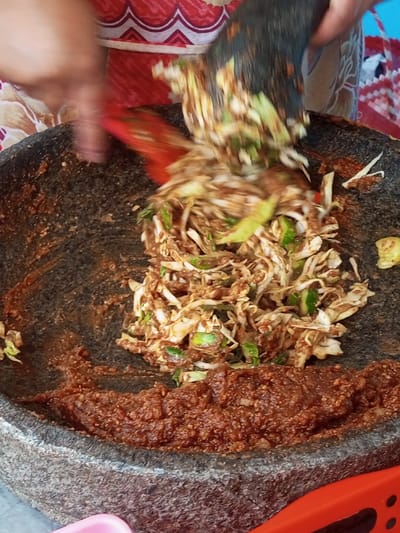 A large stone mortar contains a base of dark red, textured sauce. Chopped vegetables, including cabbage and cucumbers, are being mixed into the sauce using a dark pestle and a red spatula. The person is blending the components. A patterned red and white cloth is visible in the background, adding to the informal, homemade cooking environment.