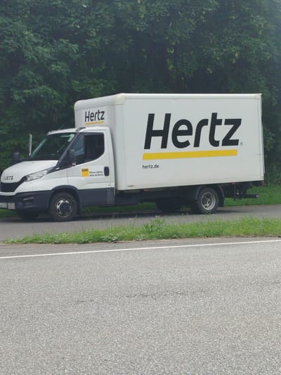A white Hertz box truck is parked on the side of a road. The truck is a Iveco model and has the Hertz logo prominently displayed in black on its side with a yellow stripe underneath. The words "Hertz" are also visible on the back of the truck. Below the Hertz logo on the cab of the truck, there is a yellow ADAC sticker with the text "Clever mieten. adac.de/hertz". The license plate of the truck is partially visible and reads "B-AX 3300". The truck is parked on a paved road with a grassy shoulder. In the background, there is a dense line of green trees. The scene appears to be set during the daytime, possibly overcast or in dappled light, as there are no strong shadows. The environment suggests a rural or suburban setting in Germany, with the presence of the ADAC sticker further indicating a European context. There are no people visible in the image. The overall impression is one of a commercial vehicle parked during its operational route.