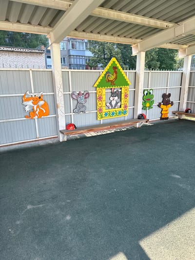 The image shows an outdoor covered area, likely part of a playground or kindergarten in Novoaltaysk, Russia. The covered structure has a corrugated metal roof supported by white beams and columns. The ground beneath is a dark green, soft-surface material. Along the back wall, which is a gray metal fence, there are colorful wooden cutouts of cartoon animals: a fox on the left, a mouse next to it, and a rabbit peeking out of a house-shaped cutout with a rooster on top. Further to the right, there is a frog and a bear dressed in yellow. Two wooden benches are positioned in front of the fence. On the left bench, there is a red toy ladybug. Another ladybug toy is on the right bench, near the edge. The background shows buildings and trees, indicating an urban or suburban setting.
