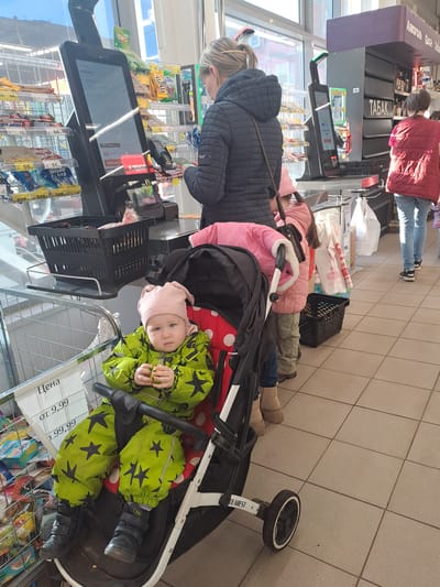 The image shows a family at a self-checkout lane in a store, likely a supermarket, based on the products visible on the shelves. The checkout station is labeled "ТАБАК," suggesting a tobacco section nearby. An individual in a black jacket is scanning items at the checkout. A young child wearing a pink hat and a star-patterned green outfit is seated in a stroller. The stroller is loaded with groceries. An older child wearing a pink jacket and beige shoes stands near the checkout area. Additional people can be seen waiting in line behind them. The location may be Elektrostal, Russia, based on the Cyrillic script visible on signage and price tags.