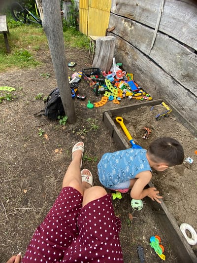 A young boy in a blue patterned shirt and grey shorts is playing in a sandbox. He is kneeling and digging in the dirt with his hands. A large yellow shovel and a blue bucket are in the sandbox with him. The sandbox is made of wood and is located outdoors. In the background, there is a pile of colorful toys, a black backpack, and a wooden fence. The ground is covered with dirt and dry leaves. A person's legs, wearing a burgundy polka dot skirt and white sandals, are visible in the foreground. The scene appears to be in a backyard in Maloarslangulovo, Russia, given the presence of a bicycle leaning against a wooden structure and the overall rustic environment.