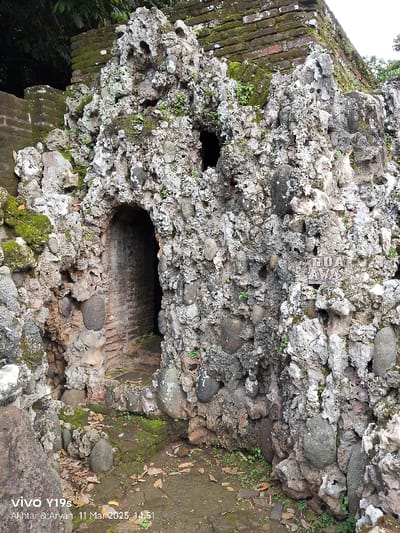 The image shows a stone wall at the entrance of a cave at the location of -6.736, 108.544 in Cirebon, Indonesia. The wall is made of large, rounded stones and has a dark, rectangular opening in the center. The cave entrance is located in a small, grassy area. The ground is covered in leaves. Above the cave entrance is a brick wall, overgrown with moss. The words "Goa Aw" are etched into the wall above the entrance, appearing to be a sign.