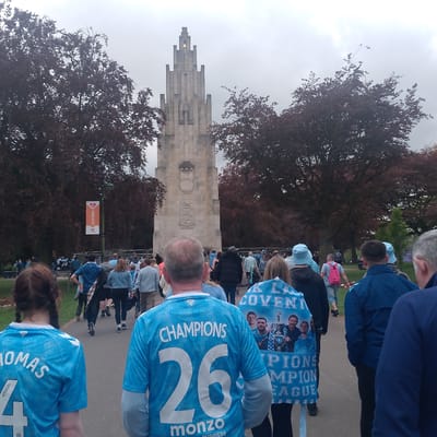 The image depicts a crowd of people walking along a pathway towards the Coventry War Memorial in Coventry, United Kingdom. The memorial is a tall, stone structure with Art Deco-inspired details. The pathway is lined with trees. The people are wearing Coventry City Football Club shirts, many of which are light blue with the team's sponsors and player names or "Champions" on the back. There is a "Love Cov" banner. The general impression is one of a public gathering or a walk to show support for the team.