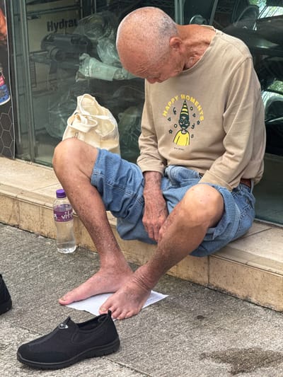 A frail, elderly man is sitting on the ground, leaning against a window with reflective glass. He is wearing a light brown long-sleeved shirt with a graphic print that reads "HAPPY MOMENTS" in yellow lettering. His lower half is clad in blue denim shorts. His skin appears weathered and shows signs of sun damage or a skin condition. His left foot is bare and rests on a piece of white paper, while a black slip-on shoe lies next to it on the concrete ground. Beside his leg is a clear plastic bottle with a purple cap, containing a light-colored liquid. A cream-colored tote bag is also visible near his legs. The glass reflection shows a blurred interior with items covered in plastic sheeting and a sign that reads "Hydrau". The scene appears to be outdoors, possibly on a sidewalk or in front of a shop. The lighting suggests it is daytime. The overall impression is one of hardship or poverty.
