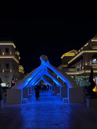 The image shows a nighttime view of the Vénitien, Qanat Quartier in Doha, Qatar. In the foreground is a sculpture illuminated with blue light. The sculpture is composed of several white arches, creating a tunnel-like effect. Underneath the arches, people are walking along a brightly lit walkway. To the right of the walkway is a blue textured wall with cube-shaped protrusions. In the background, buildings with ornate, Venetian-style architecture are visible, illuminated with warm, golden light. A dark body of water extends into the left side of the image.