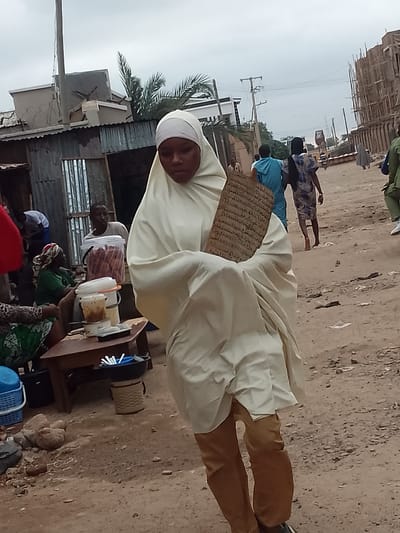 The image depicts a street scene in Pantame, Nigeria. In the foreground, a woman wearing a cream-colored hijab and matching long garment, with brown trousers visible beneath, walks towards the right of the frame. She is holding a rectangular, textured object with text inscribed on it. To her left, a low wooden table is set with several containers, some filled with food items and drinks, and a few plastic cups. Behind the table, a man in a light-colored t-shirt stands next to a large transparent container filled with what appears to be food. Further to the left, a corrugated metal structure with a small open doorway is visible, and a woman in a green top and patterned headscarf is seated. In the background, other individuals are walking along the dusty street. One person in the middle ground wears a blue garment, and another wears a patterned dress. On the right side of the background, a multi-story building under construction is prominent. The environment is characterized by a dirt road, scattered debris, and a cloudy sky.