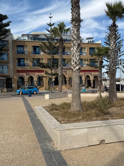A bright blue sedan is parked on a pedestrian walkway in front of a tan-colored building with blue-trimmed windows and balconies. Palm trees with rough, textured trunks are scattered throughout the scene, casting shadows on the sandy ground. The building appears to be a hotel, with "RAMADA" visible on its facade. The sky is blue with wispy clouds. In the background, there are more buildings, some with outdoor seating areas. A few people are visible, including one sitting on a bench and others standing near the hotel entrance. The scene is likely in Essaouira, Morocco, given the architectural style and presence of palm trees.