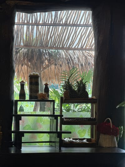 The image depicts an indoor scene with a view through a window to an outdoor tropical environment. The window frame is constructed from rough, dark wood, resembling tree trunks. Through the window, a thatched roof and lush green foliage are visible.
Inside, on a dark wooden surface, are several decorative items. On the left, a tiered wooden shelf holds three containers. The tallest, in the center, is a glass jar filled with what appears to be layered nuts or seeds, topped with small white flowers. Flanking it are two smaller, dark glass bottles with conical lids, also containing visible granular contents. A pale pink artificial flower rests on the shelf.
To the right of the first shelf, a second, lower wooden shelf displays three pineapples. Below this, another shelf has a tray containing coffee beans and a second tray with several silver spoons. To the far right, a white woven basket overflows with a dense arrangement of red flowers.
The background seen through the window shows a vibrant green garden with tropical plants. The overall ambiance suggests a serene, possibly resort or restaurant setting in a warm climate. The location provided, Nam Hoa Lư Ward, Vietnam, aligns with this tropical aesthetic.
