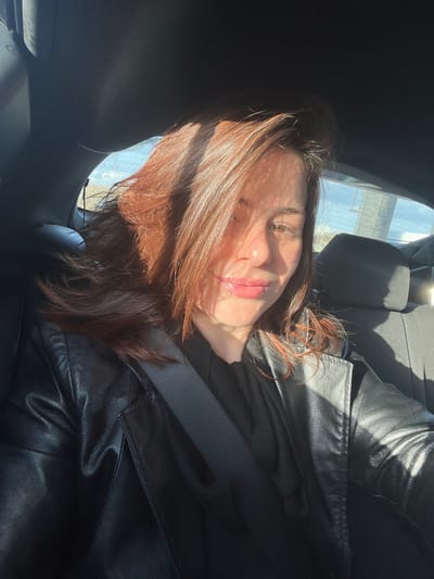 A female individual is depicted in a close-up, seated within the rear passenger compartment of a vehicle in Lille, France. She has reddish-brown hair, illuminated by direct sunlight streaming from the left, which creates distinct horizontal shadow patterns across her face. Her lips are light-colored and exhibit a subtle sheen. She is wearing a black leather jacket over a dark garment, and a black seatbelt is visible diagonally across her left shoulder and chest. The subject is looking towards the camera with a slight smile. Through the rear windows of the vehicle, a bright, clear sky is visible, along with blurred distant outdoor elements including what appears to be a utility pole and vegetation, indicating an exterior environment under strong daylight conditions.
