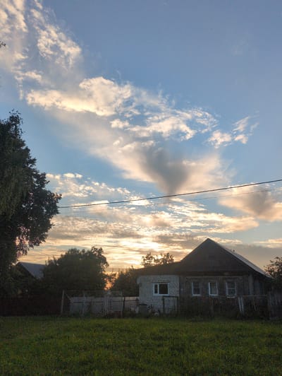 The image depicts a rural scene in Velikovo, Russia, at sunset. The sky is a prominent feature, filled with a mixture of light blue and dramatic clouds illuminated by the setting sun. Streaks of golden and orange light break through the clouds, creating a warm and serene atmosphere. A power line stretches diagonally across the sky, adding a man-made element to the natural landscape.
To the right of the frame, a rustic, two-story house is visible. The lower part of the house appears to be made of white brick, while the upper story is constructed from dark wood. Several windows are visible on the upper floor, and one window is present on the brick portion. The house is partially obscured by dense, dark green trees, which also frame the bottom and right edges of the image, suggesting a natural, somewhat wild setting. A fenced area is also discernible behind the trees, hinting at a yard or garden. The overall impression is one of a peaceful, quiet evening in the countryside. The time of day is clearly sunset, indicated by the warm lighting and the cloud formations. The weather appears clear, with the sunset creating a beautiful spectacle. There is no visible text in the image.