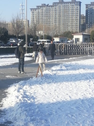 The image depicts an outdoor urban scene in Fengtai District, China, under clear skies with significant snow coverage on the ground. Several large, multi-story residential or commercial buildings dominate the background. A paved area, likely a sidewalk or pedestrian path, traverses the midground, partially covered by irregularly shaped snowdrifts.
In the immediate foreground, a young child, approximately 5-8 years old, dressed in a light-colored winter coat and pants, stands in a patch of snow. A small, brown, wooden A-frame structure is positioned upright in front of the child.
To the left, a male adult wearing a dark hooded jacket, blue jeans, and sneakers, with a dark backpack, stands facing right, looking down at a handheld device.
In the midground, a group of individuals is walking right-to-left. A male adult in a dark suit jacket and pants is prominently visible among them, walking near a line of metallic bicycle racks or barriers. Behind these barriers, a white single-story building with a red-tiled roof is visible, along with a parking area containing multiple parked cars and other pedestrians. Bare trees are present throughout the scene, consistent with a winter environment. Shadows are cast by the visible light source, indicating a sunny day.