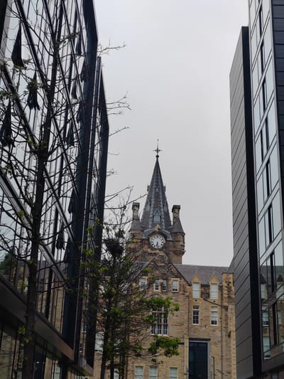 This media file captures an exterior view of a historical stone building with a prominent clock tower, nestled between two modern glass-fronted buildings. The historical structure features a steep, grey, slate-tiled roof and numerous windows with white frames, some of which appear to be of varying sizes and shapes. The clock face on the tower is clearly visible, with Roman numerals. A weather vane tops the clock tower.
The scene is framed by the reflective surfaces of the contemporary buildings, which create a striking contrast with the aged architecture. Bare-branched trees, some with nascent green leaves, are positioned in the foreground and mid-ground, suggesting a temperate season, likely spring or early summer. One tree on the left has a complex network of branches, while another on the right displays a large bird's nest.
The sky is overcast and a pale grey, indicating a cloudy day, and the overall lighting is diffused, suggesting it is likely daytime, though no specific time can be definitively determined from the light alone. The reflections in the modern buildings show glimpses of the sky and the surrounding urban environment. The image appears to be taken from a street level perspective, looking upwards towards the buildings. The architectural styles present suggest an urban setting where old and new developments coexist, possibly in a city center. No people are visible in the frame. No text is discernible in the image.