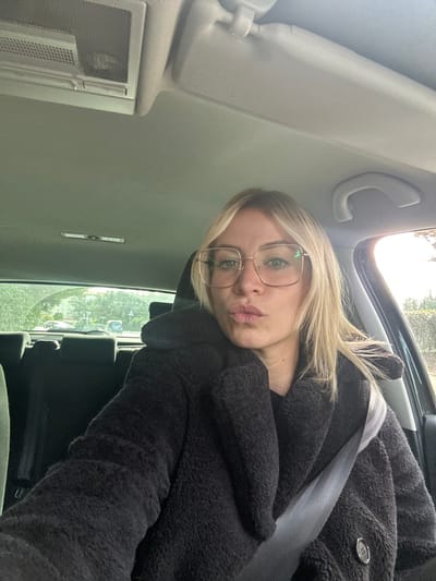 A blonde woman, appearing to be in her 20s or 30s, is seated in the front passenger seat of a vehicle, looking directly towards the camera with a pursed-lip expression. She wears large, square-framed, light-colored eyeglasses and a thick, dark, textured coat over a black top. A grey seatbelt is fastened across her chest and shoulder. The car's light-colored headliner and an overhead console are visible above her. Through the vehicle's windows, a street scene in Antibes, France, is visible under ambient daylight, featuring trees, abundant greenery, and distant buildings. Faint indications of other vehicles or street infrastructure are discernible in the background.