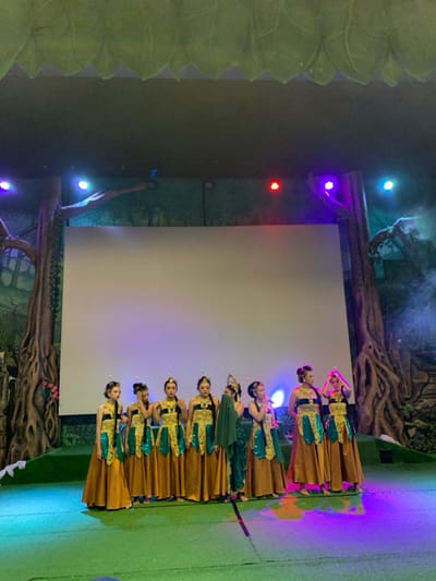 Nine young women are standing on a stage in front of a large white screen. They are wearing gold and green traditional Indonesian costumes and have their hair elaborately styled with flowers.  They are all looking at the camera, with some of them smiling. The stage is decorated with fake trees and greenery, and the lighting is dim. The stage is lit with purple and blue lights. This performance is taking place in a theater in Batu, Indonesia at the coordinates -7.885 latitude and 112.524 longitude.