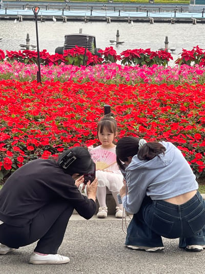 The image depicts a woman in black clothing taking a photograph of a young girl in a white dress and another woman in blue clothing, all standing in front of a wall of red and pink flowers. The location is in Guang Zhou Shi, China, at the coordinates 23.115 latitude and 113.319 longitude. The woman in black clothing is holding a smartphone in her right hand and looking at the screen. The girl is holding a white fan in her left hand and looking at the camera. The woman in blue clothing has her back to the camera, is looking at the girl, and is wearing blue jeans and a light blue sweater. The background is a wall of red and pink flowers.