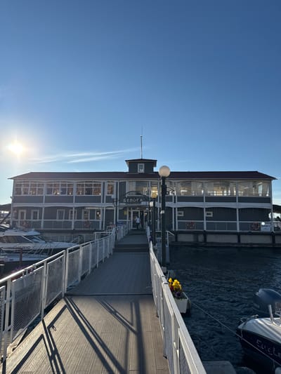 The image shows a bright, clear day with a deep blue sky and a low sun casting long shadows, suggesting it is late afternoon or early morning. The scene is a waterfront with a multi-story building that appears to be a restaurant or establishment on a river or canal. The building has a dark roof, gray siding, and many large windows with sheer white curtains. White railings and a wooden walkway lead towards the building.
To the right of the building, a boat with a Yamaha engine is docked. A white planter with yellow and purple flowers is visible on the walkway near the boat. The walkway itself is made of wooden planks, and there are metal stairs leading down to the water. A few life preservers are attached to the building.
Visible text on the building includes "БЕЛЫЙ ДОМ" and "РЕСТОРАН". There are also some Cyrillic characters on the boat. The overall atmosphere is calm and serene, with the bright sunlight creating a pleasant ambiance. No people are clearly visible, but the setting suggests a place of public gathering or dining. The location is identified as Novokuybyshevsk, Russia.