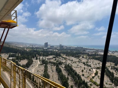 An aerial, wide-angle shot captures a sprawling cemetery in Haifa, Israel, situated on a hillside overlooking a modern city with high-rise buildings and the Mediterranean Sea. The foreground is partially obstructed by a yellow metal railing and a dark diagonal pole, suggesting the perspective is from a high vantage point, possibly a construction site or observation deck. The cemetery itself is densely packed with tombstones, interspersed with numerous cypress trees, creating a textured, dark green and light grey landscape. Winding paths navigate through the graves. In the midground, the city's architecture, including several tall buildings, is visible. The background features the distant coastline and the expansive, blue sea under a sky with scattered white clouds. The overall scene depicts a juxtaposition of the eternal repose of the cemetery with the dynamic, modern city and the natural environment.