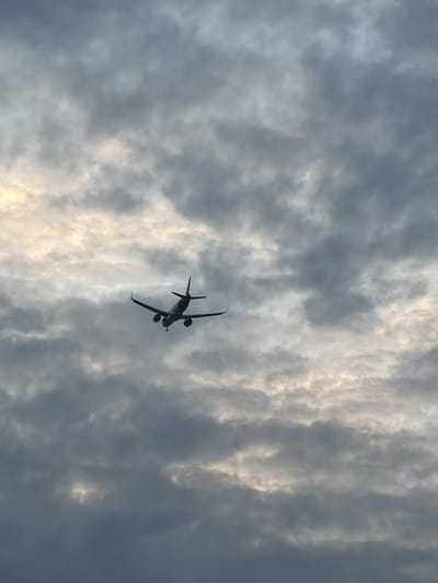 A silhouette of a passenger airplane is captured mid-flight against a cloudy sky. The aircraft, seen from below and slightly from the rear, has its landing gear down, suggesting it is in the process of landing or taking off. The sky is filled with layers of grey and white clouds, with patches of brighter light suggesting the presence of the sun filtering through. The image does not provide any discernible information to suggest a specific location in Delhi, India.