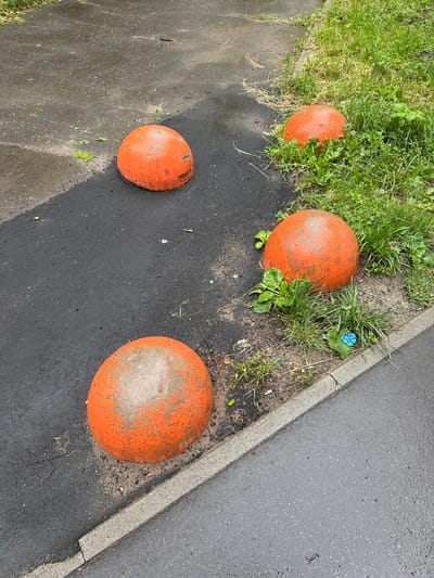 The image depicts four orange concrete bollards positioned along the edge of an asphalt pathway and a grassy area. The bollards are weathered, with some of the orange paint chipped away, revealing the gray concrete beneath. The pathway appears wet, suggesting recent rain. There are patches of grass and some small green plants growing around the bollards and along the edge of the pathway. In the distance, more greenery is visible. The scene is outdoors, with a neutral lighting. There are no people or animals visible in the image. The location is identified as Vologda, Russia.
