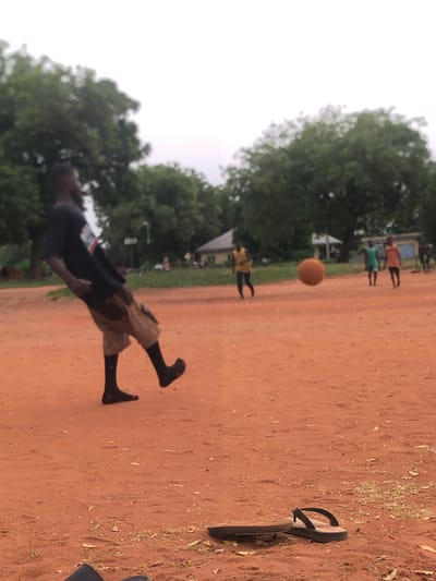 A dynamic outdoor photograph captures a soccer game in progress on a reddish-brown dirt field in Nsugbe, Nigeria. The scene is dominated by the earthy tones of the unpaved ground, contrasting with the lush green foliage of trees in the background and a bright, overcast sky above.

In the foreground, a young person, likely male, is captured in mid-action, seemingly having just kicked or about to kick an orange ball. The ball is suspended in the air to their right. The player wears a dark short-sleeved top with a blurry, indistinct graphic or text on the front, and light-colored khaki shorts. Their legs are bent and active, suggesting a powerful movement, and they appear to be barefoot on the dusty ground.

Scattered on the reddish-brown field, which has sparse vegetation and fallen leaves, a pair of black flip-flops can be seen to the right, implying they were shed for the game. In the mid-ground and background, several other figures, likely children or young adults, are visible on the field, some wearing brightly colored tops (yellow, green, orange). Their presence confirms a group activity.

The time of day appears to be daytime, with soft, diffused lighting suggesting an overcast sky. The natural setting, combined with the activity, evokes a sense of energetic play in a community outdoor space. No clear, legible text is visible other than the indistinct graphic on the player's shirt.