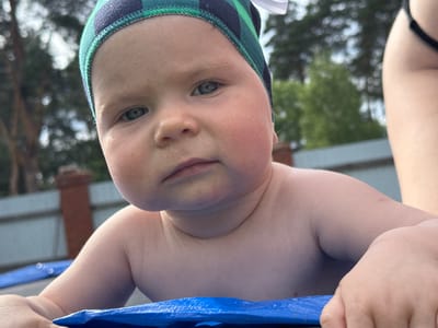 A close-up shot shows a baby wearing a green and navy checkered hat. The baby is looking directly at the camera with a neutral expression. The background is blurred, showing trees and a fence, suggesting an outdoor setting during the daytime. The baby appears to be leaning on a blue surface, possibly a mat or trampoline. The lighting and soft focus suggest a natural, casual moment captured outdoors. The overall impression is one of a candid portrait of a young child.