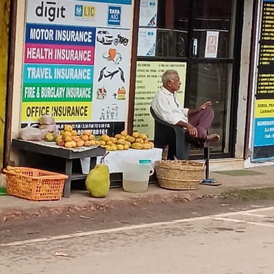 An elderly man is sitting in a black chair with a black frame, resting his bare foot on a black stand in front of a building on a street in Oxel, India. He is wearing a light-colored long-sleeved shirt and maroon pants. His hair is gray and receding. The building is next to a sidewalk. To the man's left is a makeshift stand displaying fruit, some of which are contained in baskets. Behind the fruit stand is an advertising board with insurance information for companies like Digit, LIC, and Tata AIG. There is another sign on the right side, but it is mostly cut off in the frame.